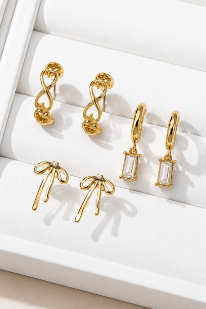 A set of six gold-toned earrings displayed on a white jewelry box. The collection includes two twisted infinity hoop earrings, two dangling earrings with rectangular clear stones, and two bow-shaped earrings with small clear stones, all arranged on a white padded tray.