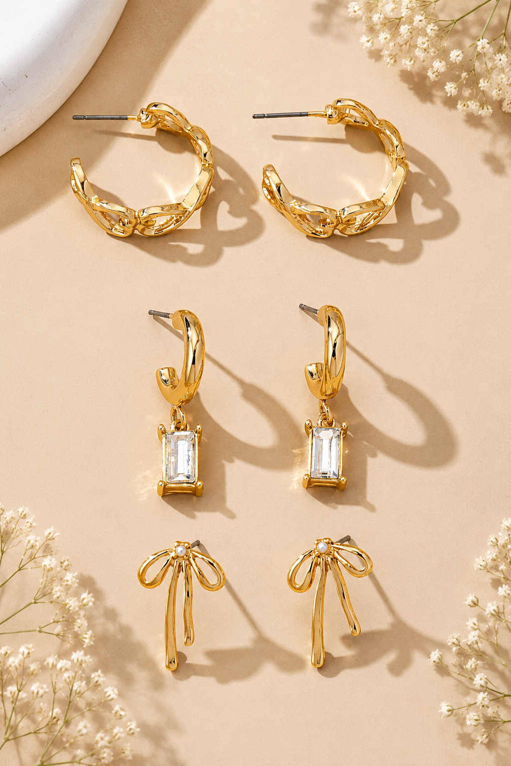 Three pairs of gold hoop-style earrings arranged on a beige background with small white floral props. The top pair is a twisted open hoop, the middle pair is a huggie hoop with a clear rectangular gemstone drop, and the bottom pair is a bow-shaped hoop.