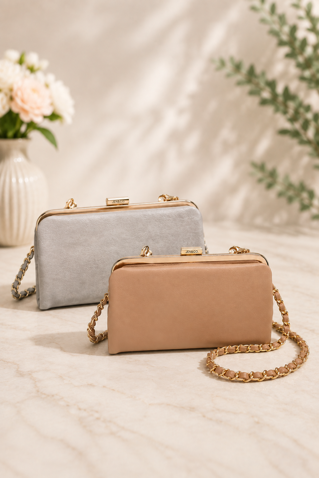 Two elegant clutch bags with gold-tone chain straps, displayed on a light marble surface. The front bag is a warm tan/beige color, and the bag behind it is a light gray color.