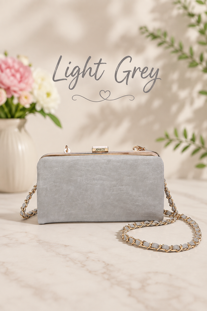 A light grey textured clutch bag with a gold-tone kiss-lock clasp and a gold chain strap, displayed on a marble surface with a floral vase in the background and the text 'Light Grey' written above.