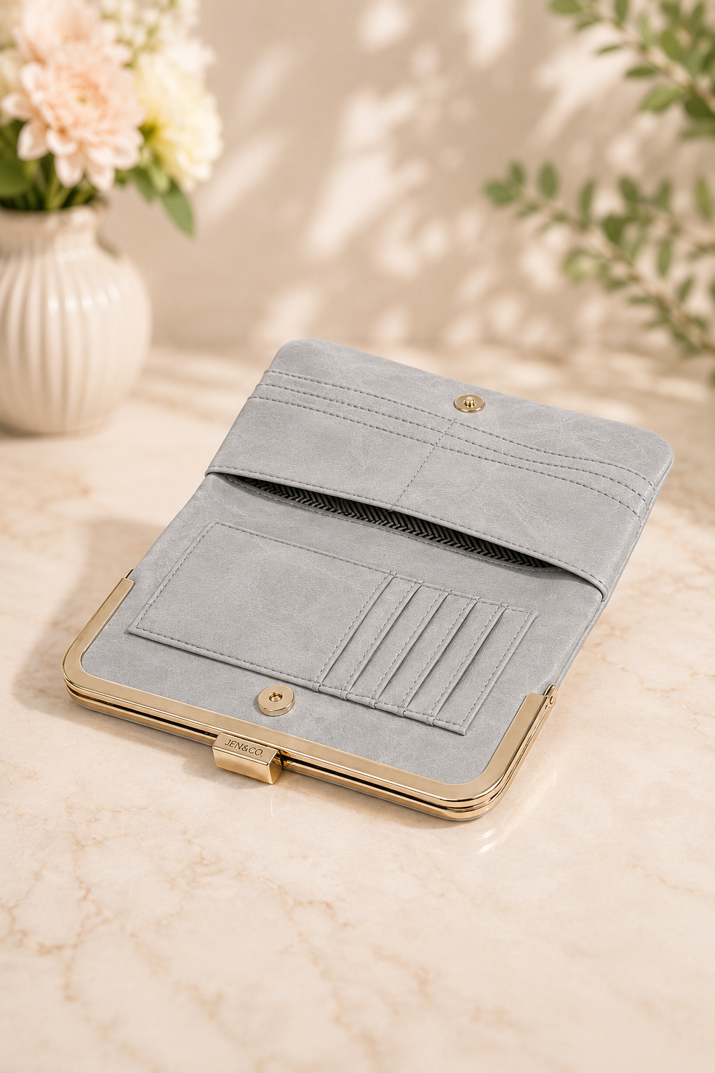 A light gray suede kiss-lock clutch wallet with gold-tone metal trim, shown open to reveal card slots and an interior pocket, set on a marble surface with soft floral background props.