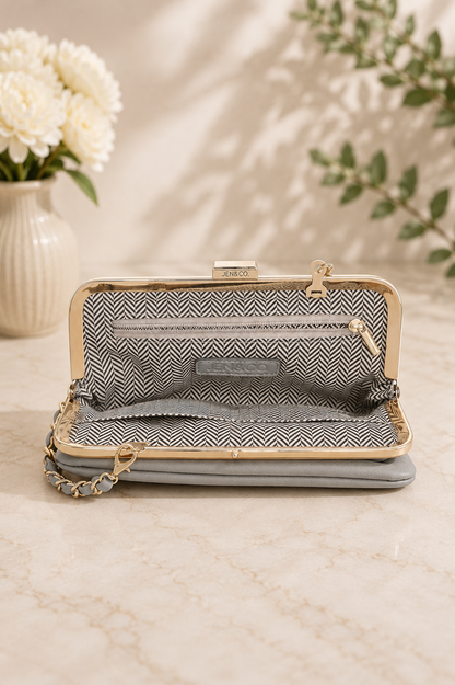 An open clutch handbag with a gray and white herringbone pattern interior, a light gray exterior, and a gold-tone metal frame, displayed on a marble surface with a vase of white flowers in the background.