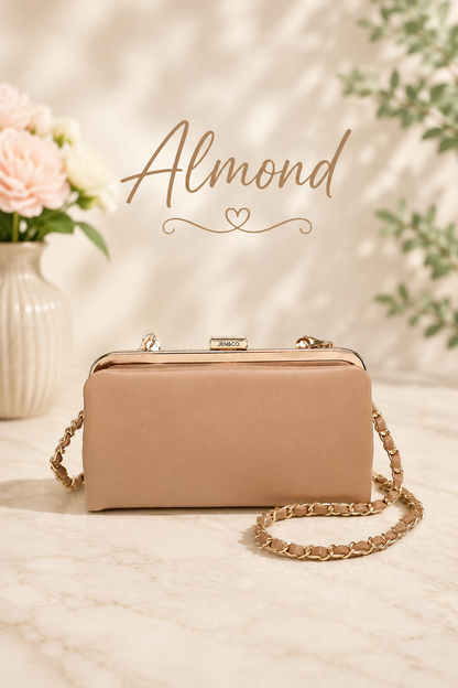 An almond beige leather kiss-lock handbag with a gold-tone chain strap, displayed on a marble surface with a vase of flowers and soft shadows in the background. The word 'Almond' is written in gold script above the bag.