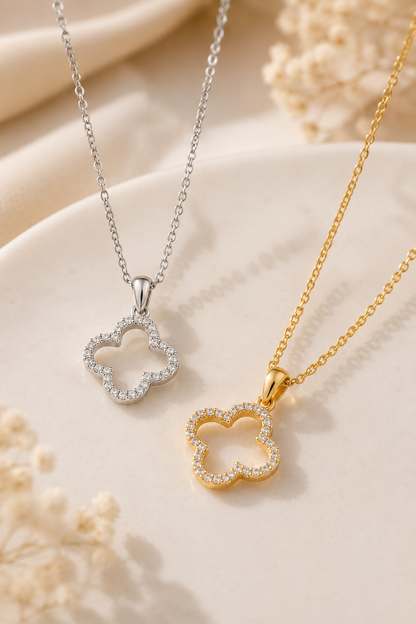 Two delicate clover-shaped necklaces with pavé-set clear stones, one in silver and one in yellow gold, displayed on a soft cream fabric background with dried floral accents.