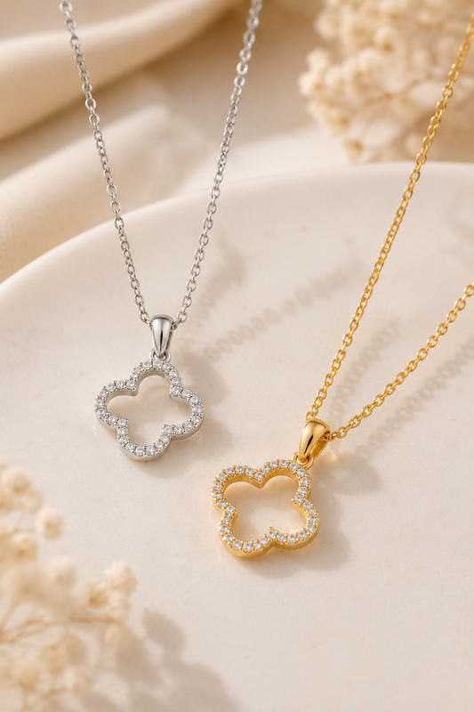 Two delicate clover-shaped necklaces with pavé-set clear stones, one in silver and one in yellow gold, displayed on a soft cream fabric background with dried floral accents.