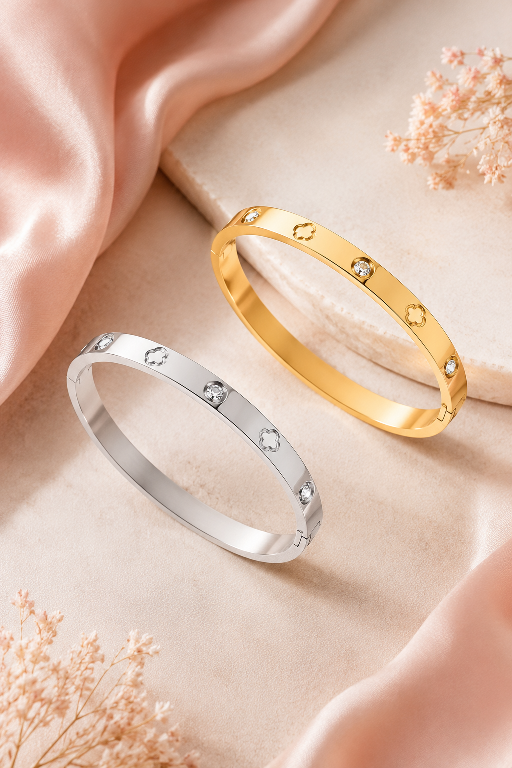Two slim bangle bracelets, one in yellow gold and one in silver, each accented with small clear stones, displayed on a soft beige fabric surface with draped pink satin and dried flowers.