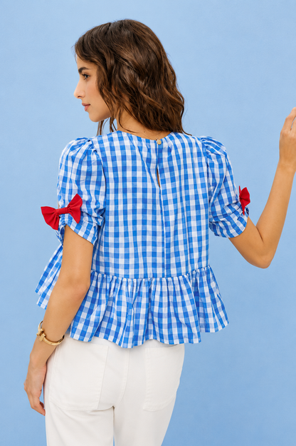 Woman wearing SO ME blue gingham ribbon detail short sleeve blouse featuring bow accents, peplum silhouette, and lightweight summer styling. Back View