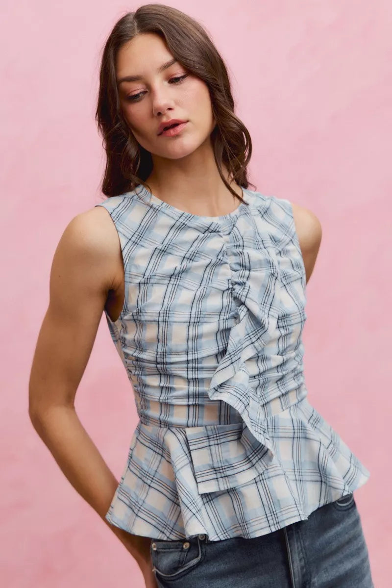 Woman wearing SO ME blue plaid sleeveless peplum top with ruffle front detail, shirred waist, and lightweight cotton fabric. Front view