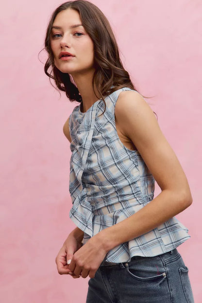 Woman wearing SO ME blue plaid sleeveless peplum top with ruffle front detail, shirred waist, and lightweight cotton fabric. Side View