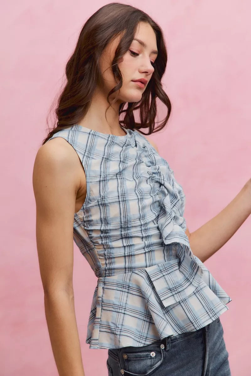 Woman wearing SO ME blue plaid sleeveless peplum top with ruffle front detail, shirred waist, and lightweight cotton fabric. right Side View