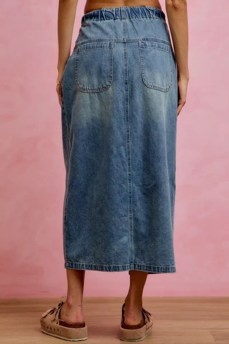 Woman wearing SO ME high waist button front denim maxi skirt with drawstring waistband, pleated waist detail, distressed wash, and front slit. Back View