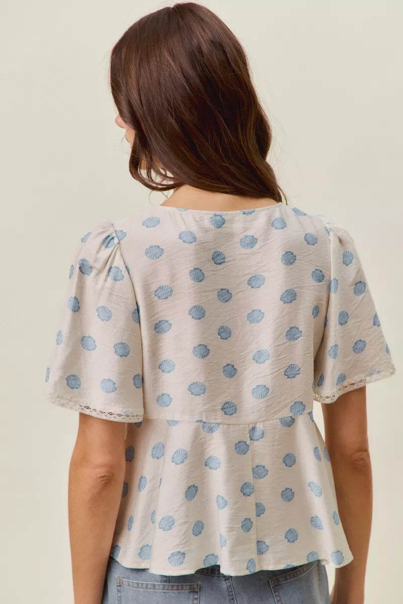 Woman wearing SO ME ivory blouse with blue shell print, tie-front neckline, flutter sleeves, and lace trim detail. Back View