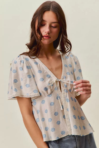 Woman wearing SO ME ivory blouse with blue shell print, tie-front neckline, flutter sleeves, and lace trim detail. Front View