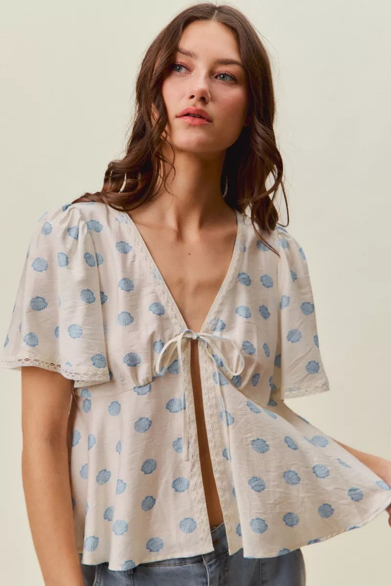 Woman wearing SO ME ivory blouse with blue shell print, tie-front neckline, flutter sleeves, and lace trim detail. Front View 2