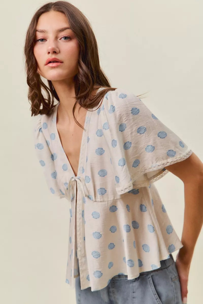 Woman wearing SO ME ivory blouse with blue shell print, tie-front neckline, flutter sleeves, and lace trim detail. Side View 2