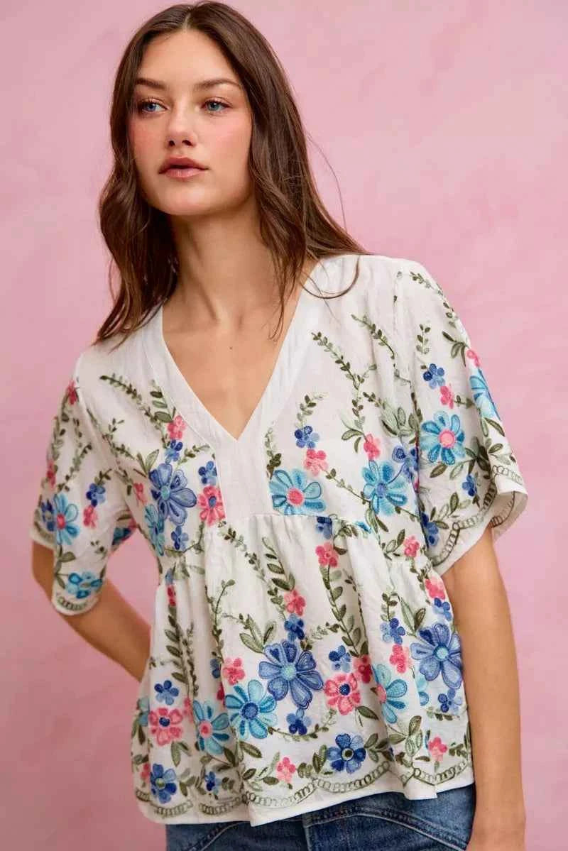 Woman wearing SO ME off white floral embroidered babydoll blouse top featuring colorful floral embroidery, flowy silhouette, short sleeves, and feminine everyday style. 
Front View