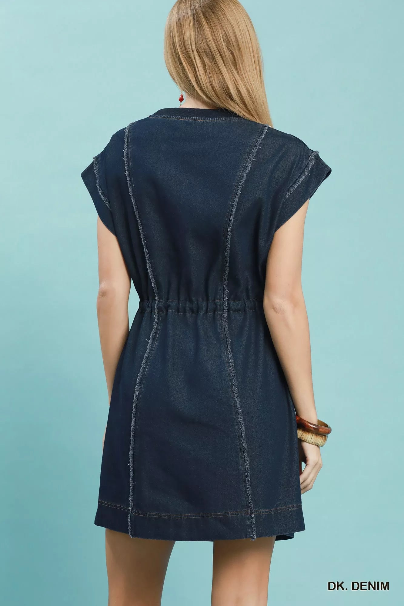 Woman wearing Umgee dark denim mini dress with raw edge seam detailing, drawstring waist, cap sleeves, and side pockets. Back View