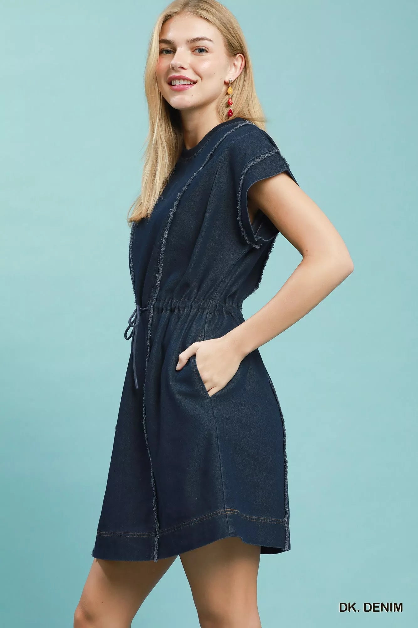 Woman wearing Umgee dark denim mini dress with raw edge seam detailing, drawstring waist, cap sleeves, and side pockets. Side View