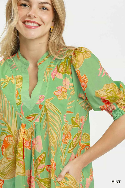 Woman wearing Umgee mint tropical floral dress with smocked sleeves, split V-neck, pockets, and flowy silhouette perfect for summer vacations and brunch. Close View