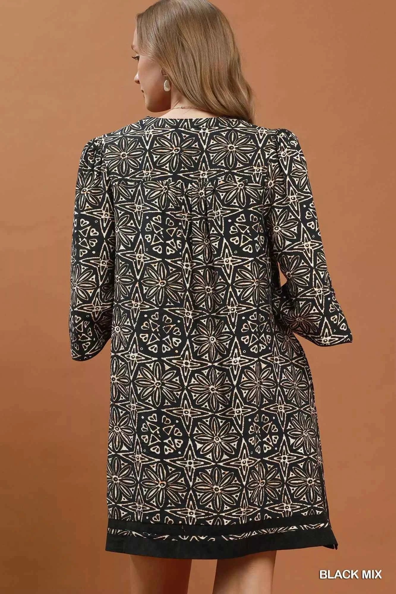 Woman wearing a black geometric print tunic dress by Umgee featuring a V-neck tassel tie, split sleeves, side pockets, and a relaxed silhouette. back view