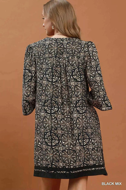 Woman wearing a black geometric print tunic dress by Umgee featuring a V-neck tassel tie, split sleeves, side pockets, and a relaxed silhouette. back view