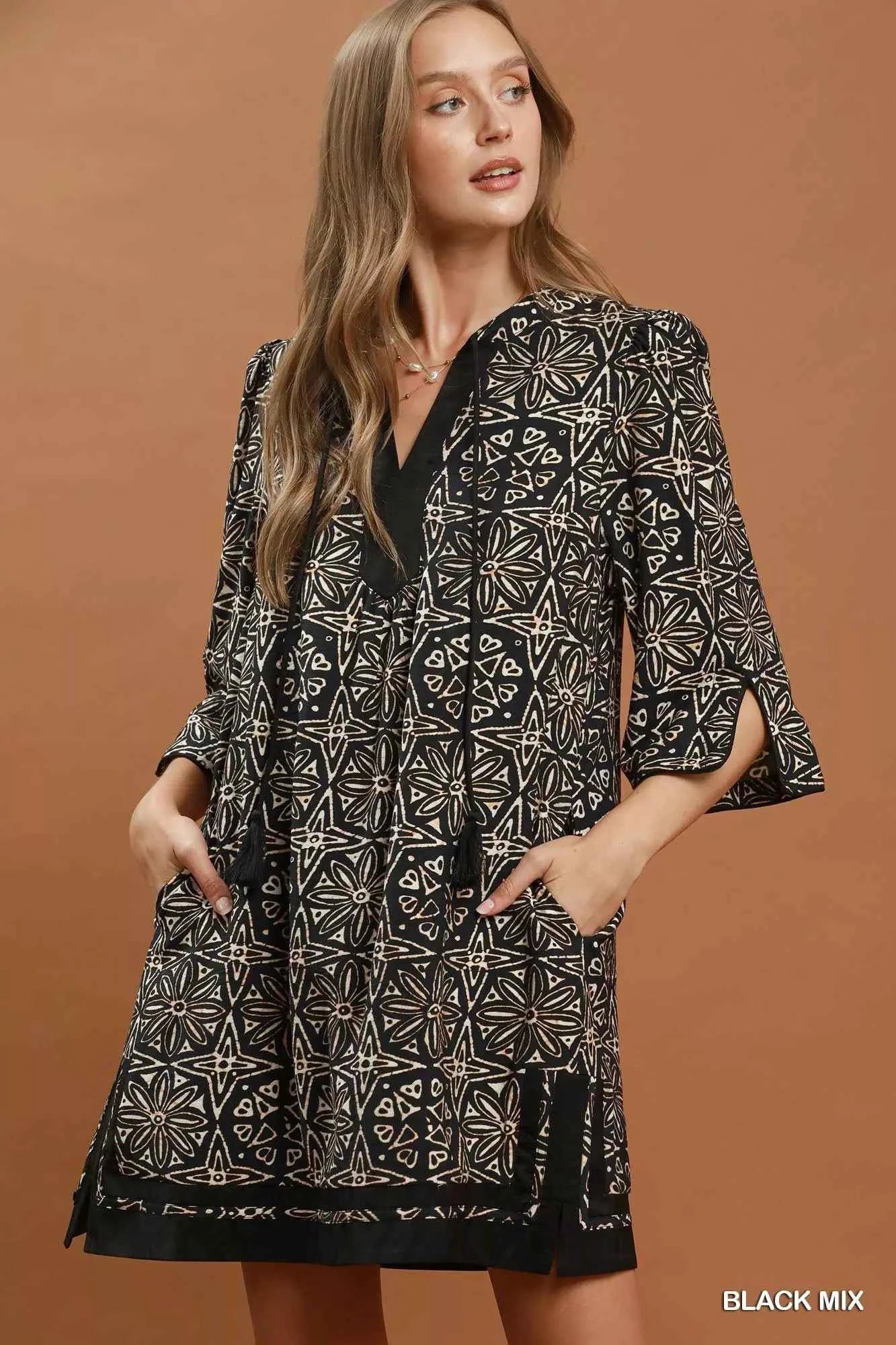 Woman wearing a black geometric print tunic dress by Umgee featuring a V-neck tassel tie, split sleeves, side pockets, and a relaxed silhouette. front view