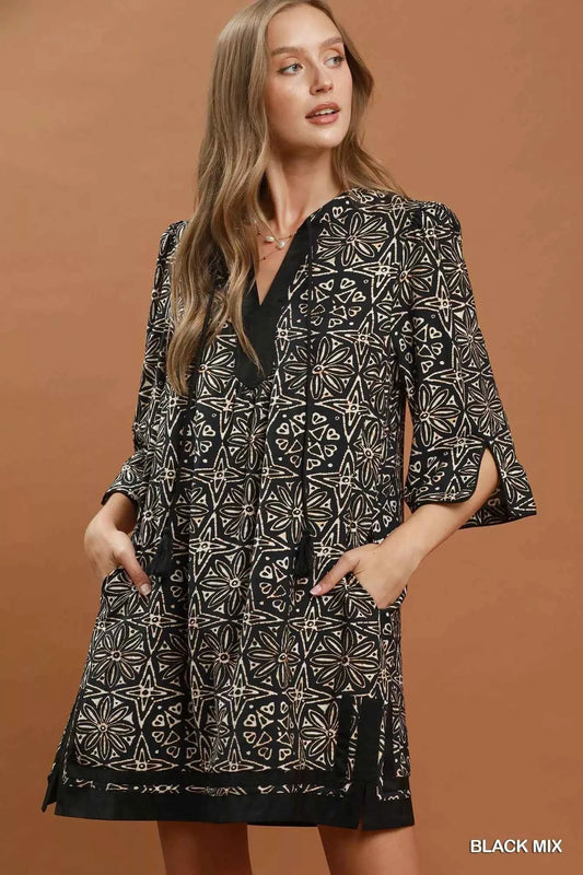 Woman wearing a black geometric print tunic dress by Umgee featuring a V-neck tassel tie, split sleeves, side pockets, and a relaxed silhouette. front view