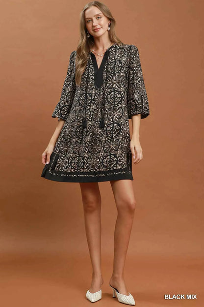 Woman wearing a black geometric print tunic dress by Umgee featuring a V-neck tassel tie, split sleeves, side pockets, and a relaxed silhouette. full view