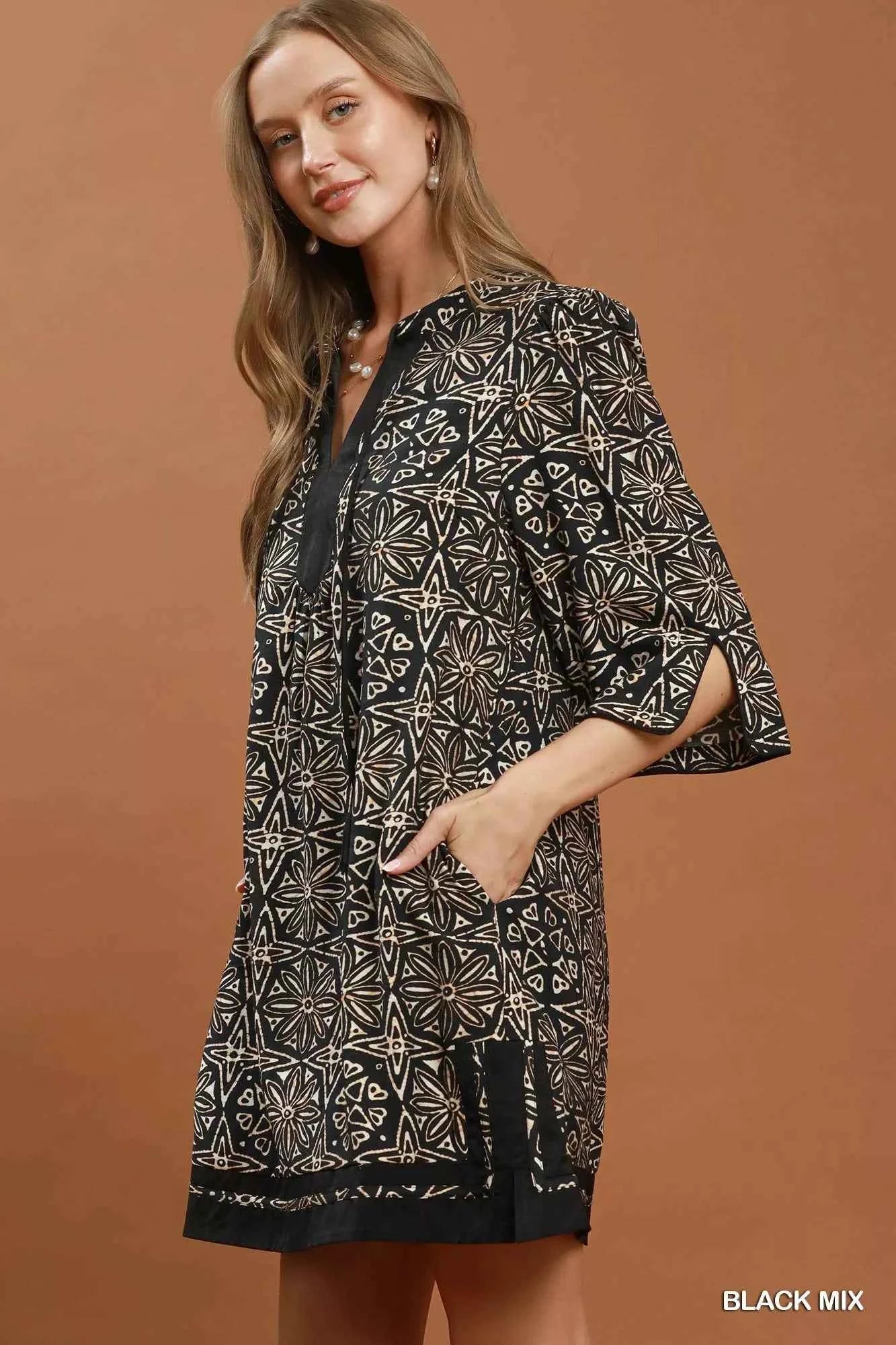 Woman wearing a black geometric print tunic dress by Umgee featuring a V-neck tassel tie, split sleeves, side pockets, and a relaxed silhouette. side view