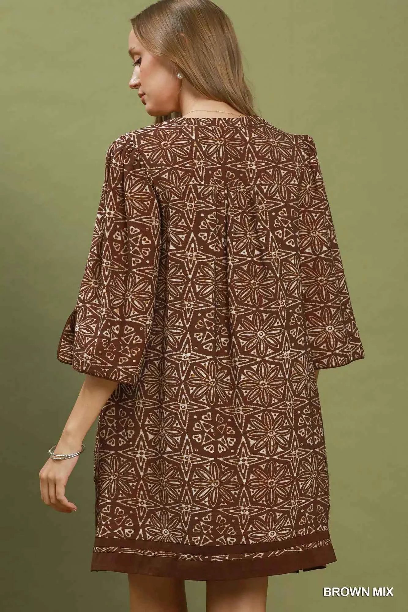 Woman wearing a brown geometric print tunic dress with a V-neckline, tassel ties, and three-quarter slit sleeves by Umgee. Back View