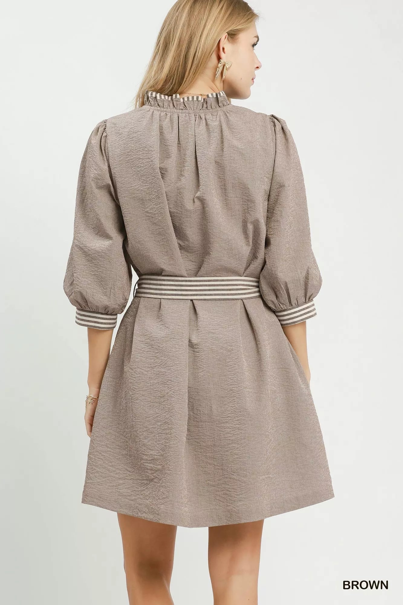 Woman wearing a brown textured mini dress by Umgee featuring striped contrast trim, an adjustable tie waist, lined construction, long balloon sleeves, and side pockets. back view