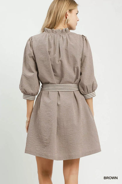 Woman wearing a brown textured mini dress by Umgee featuring striped contrast trim, an adjustable tie waist, lined construction, long balloon sleeves, and side pockets. back view