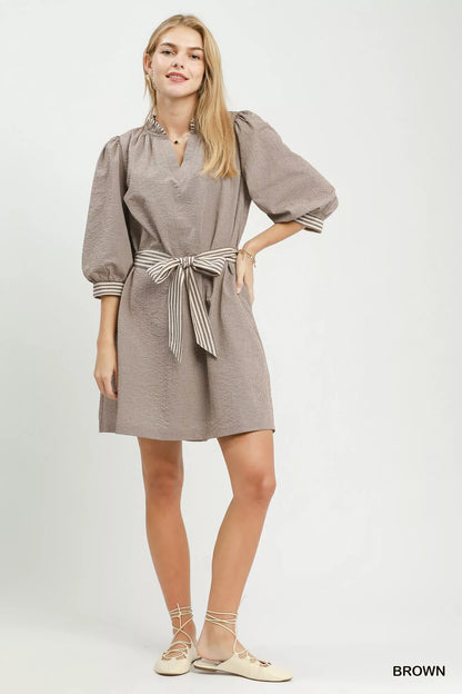 Woman wearing a brown textured mini dress by Umgee featuring striped contrast trim, an adjustable tie waist, lined construction, long balloon sleeves, and side pockets. full view