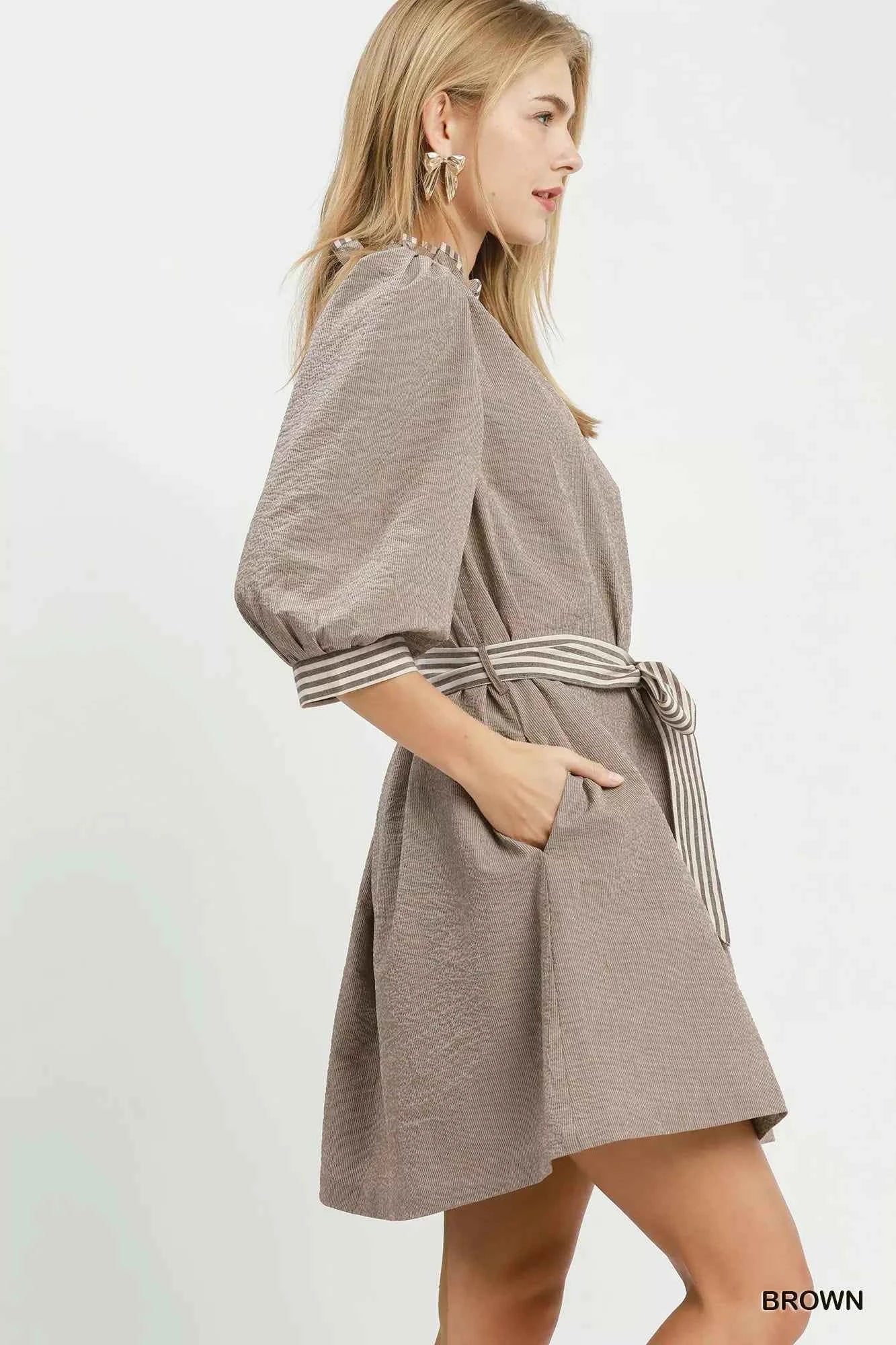 Woman wearing a brown textured mini dress by Umgee featuring striped contrast trim, an adjustable tie waist, lined construction, long balloon sleeves, and side pockets. side view
