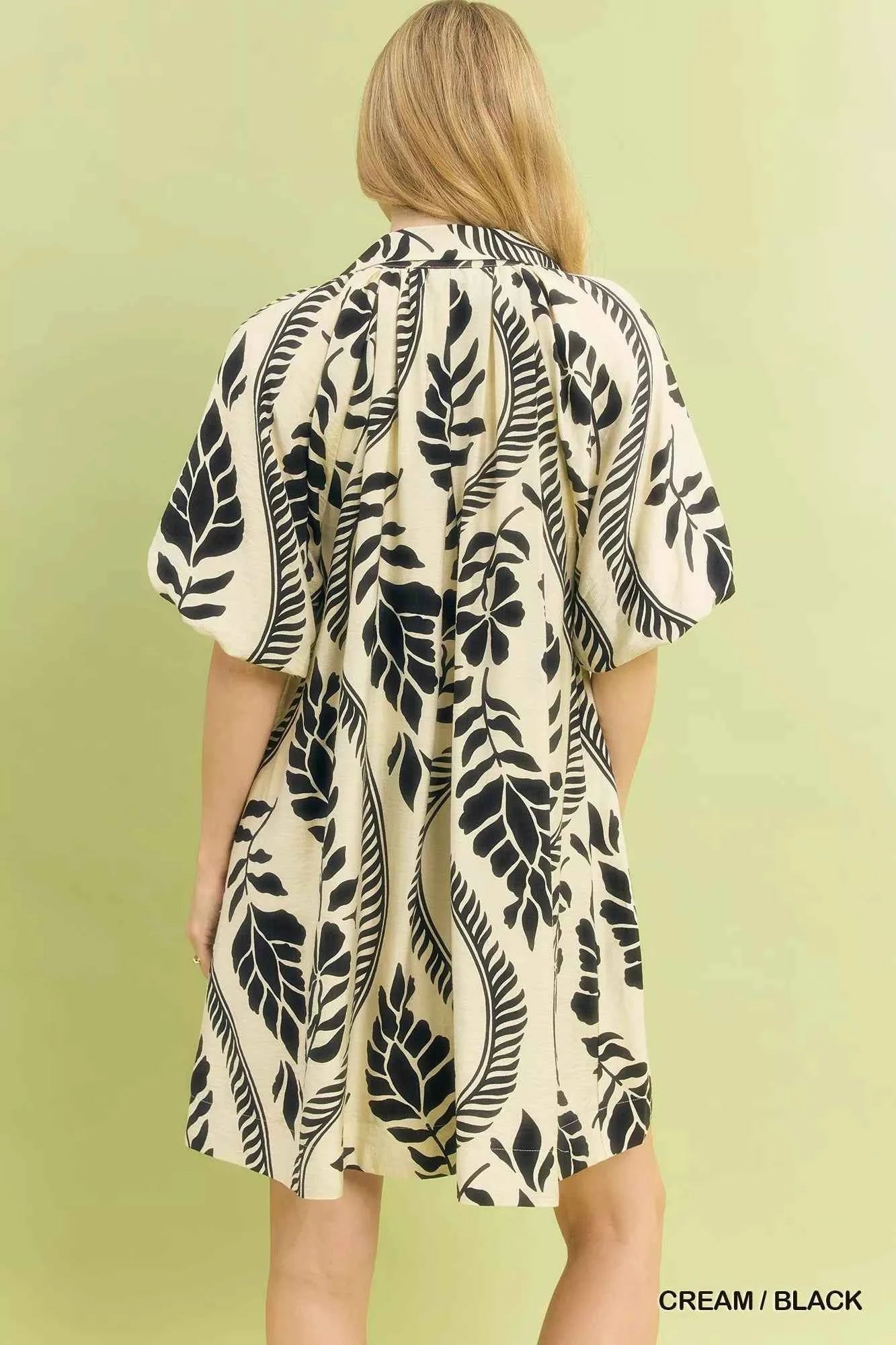 Woman wearing a cream and black leafy ogee print mini dress with a collared V-neckline and side pockets by Umgee. Back View