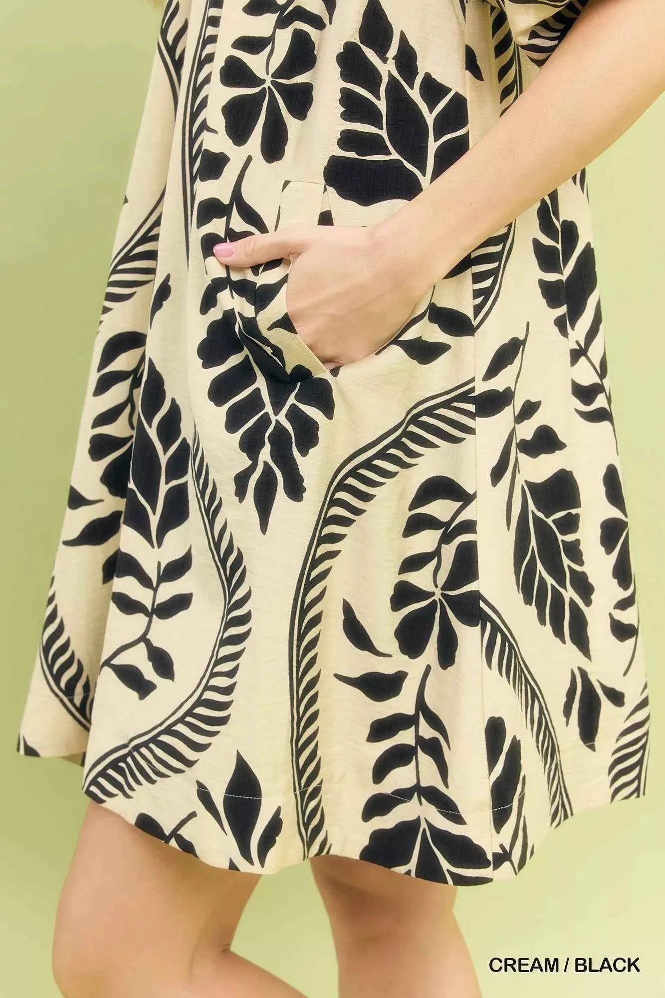 Woman wearing a cream and black leafy ogee print mini dress with a collared V-neckline and side pockets by Umgee. close View