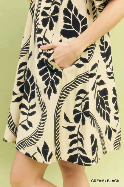 Woman wearing a cream and black leafy ogee print mini dress with a collared V-neckline and side pockets by Umgee. close View