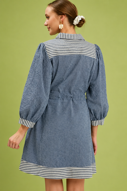 Woman wearing a denim blue belted mini dress by Umgee featuring striped contrast trim, puff sleeves, a collared neckline, removable waist tie, and side pockets. back view