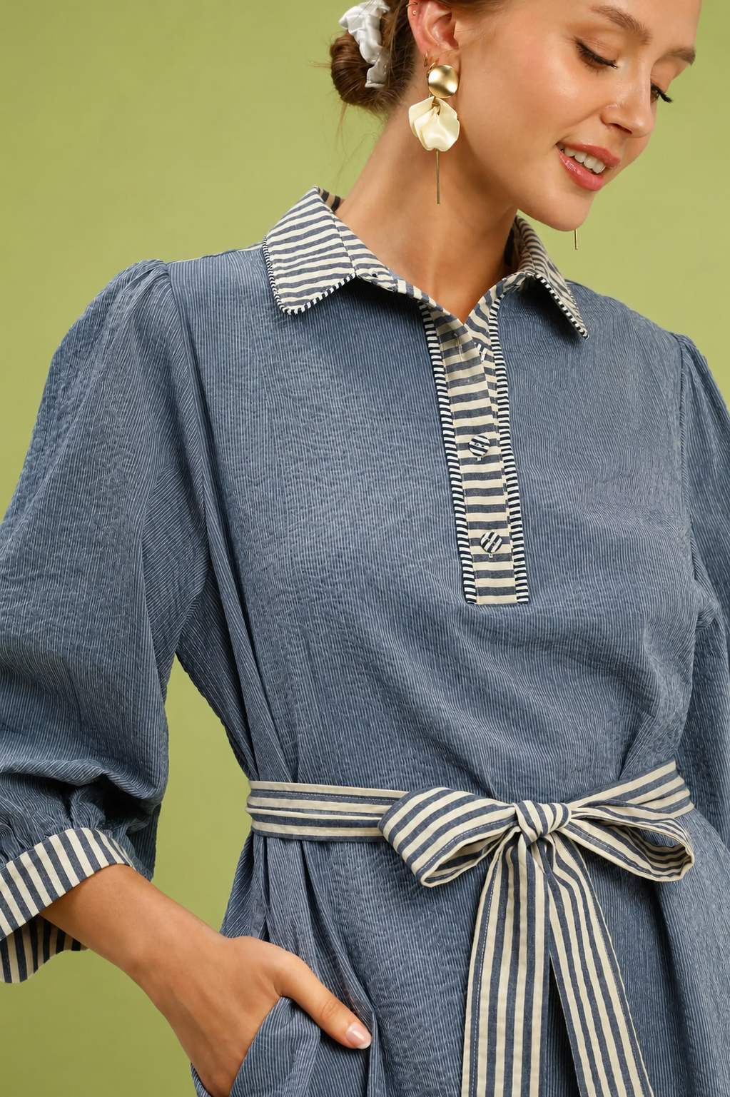 Woman wearing a denim blue belted mini dress by Umgee featuring striped contrast trim, puff sleeves, a collared neckline, removable waist tie, and side pockets. close view