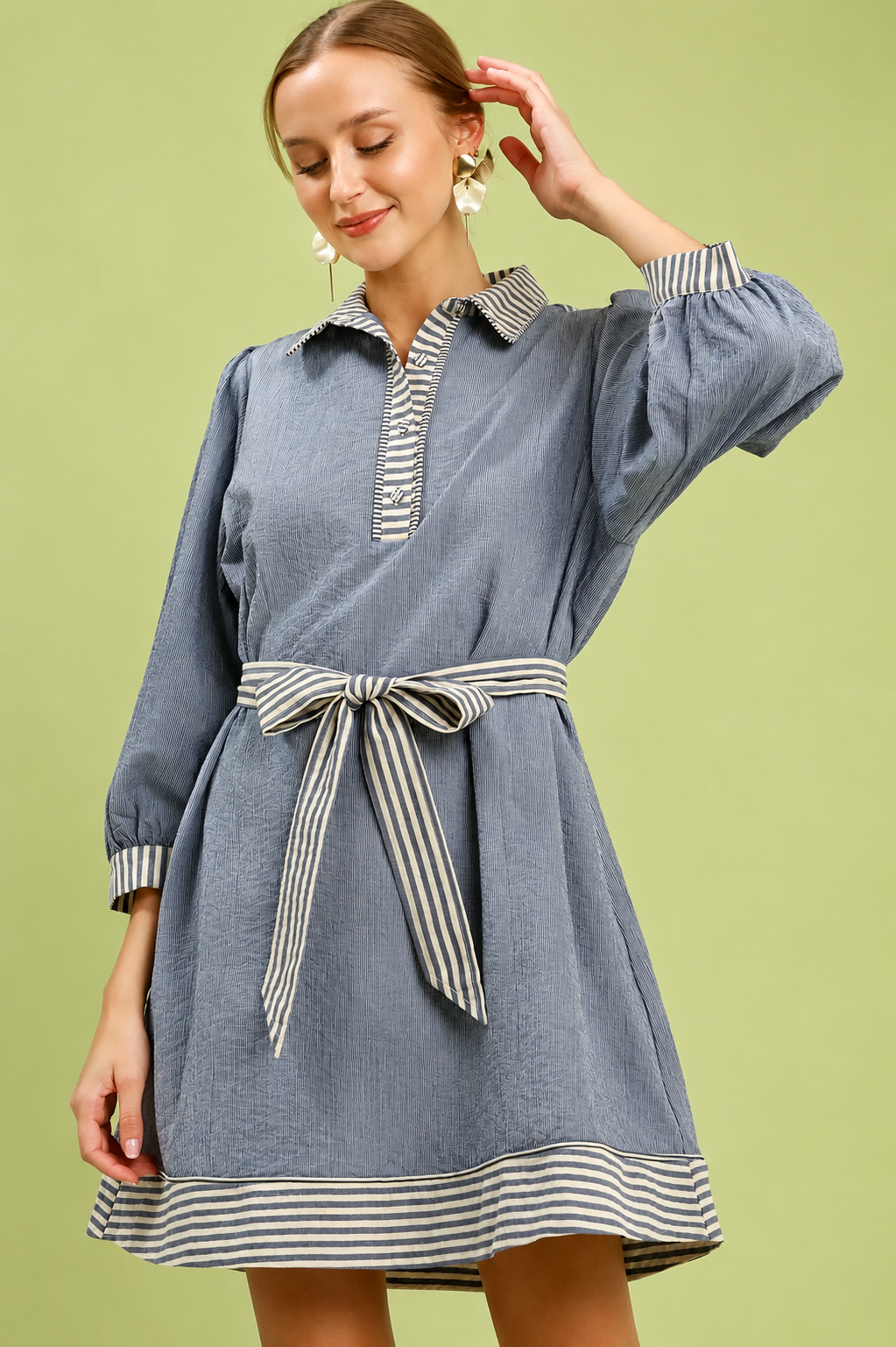 Woman wearing a denim blue belted mini dress by Umgee featuring striped contrast trim, puff sleeves, a collared neckline, removable waist tie, and side pockets. front view