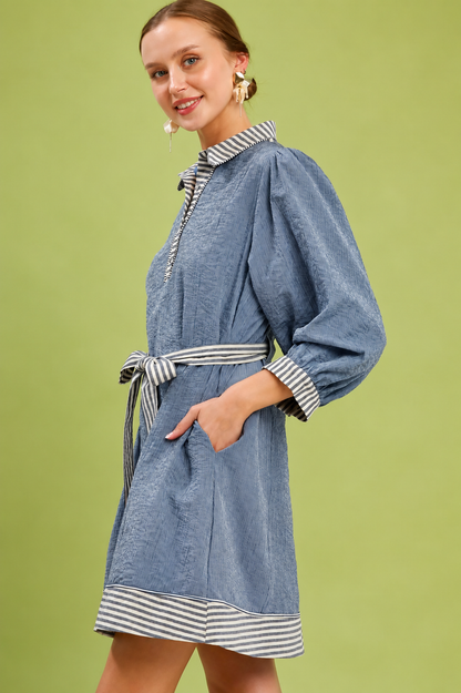 Woman wearing a denim blue belted mini dress by Umgee featuring striped contrast trim, puff sleeves, a collared neckline, removable waist tie, and side pockets. side view