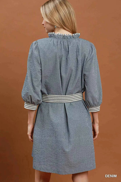 Woman wearing a denim blue textured mini dress with contrast striped trim, puff sleeves, side pockets, and a tie waist by Umgee. back view