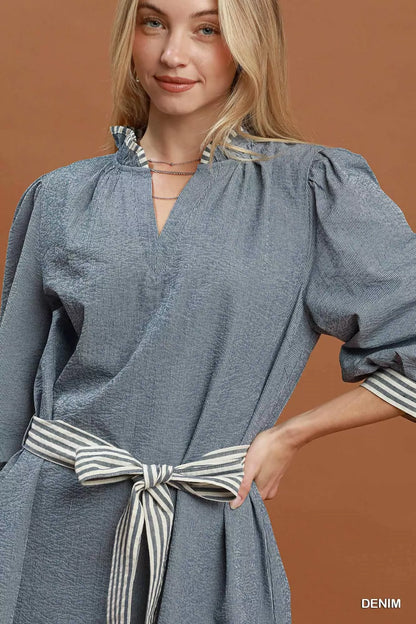 Woman wearing a denim blue textured mini dress with contrast striped trim, puff sleeves, side pockets, and a tie waist by Umgee. close view