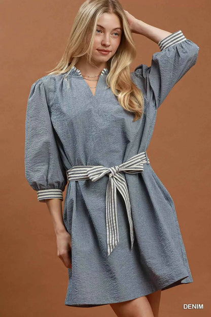 Woman wearing a denim blue textured mini dress with contrast striped trim, puff sleeves, side pockets, and a tie waist by Umgee. front view belted