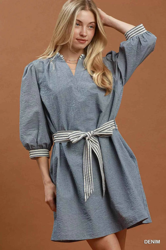 Woman wearing a denim blue textured mini dress with contrast striped trim, puff sleeves, side pockets, and a tie waist by Umgee. front view belted