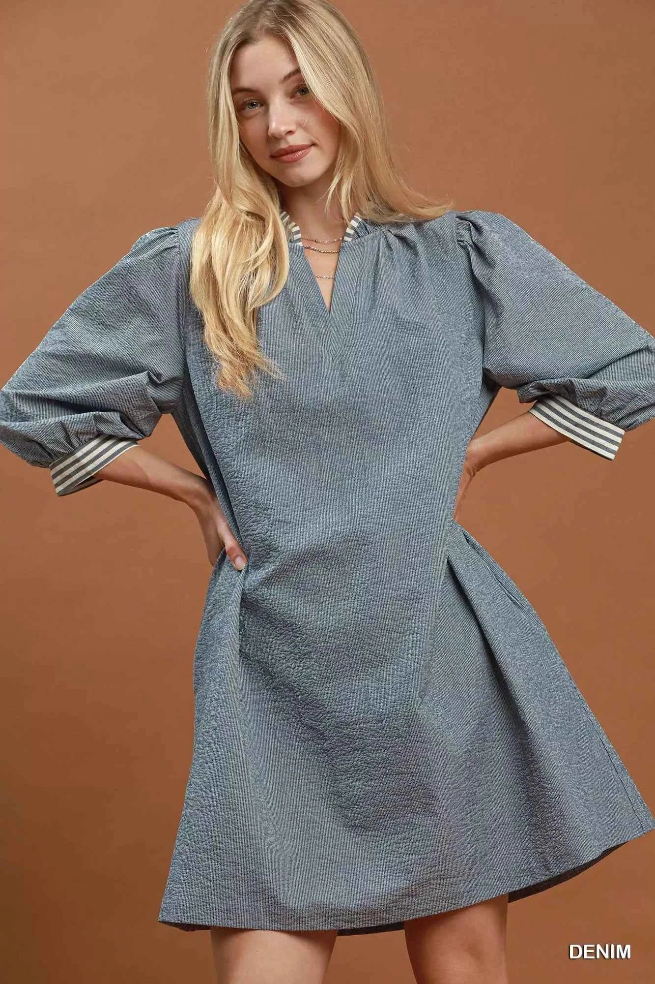 Woman wearing a denim blue textured mini dress with contrast striped trim, puff sleeves, side pockets, and a tie waist by Umgee. front view unbelted