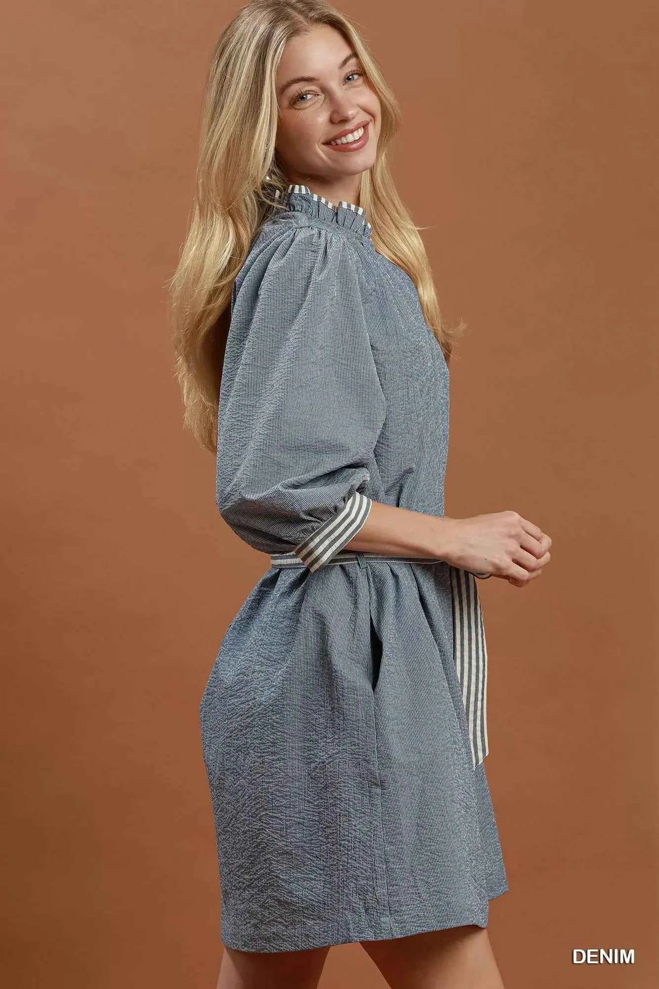 Woman wearing a denim blue textured mini dress with contrast striped trim, puff sleeves, side pockets, and a tie waist by Umgee. side view