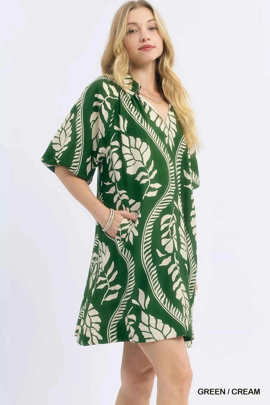 Woman wearing a green and cream leafy ogee print mini dress with a collared V-neckline and side pockets by Umgee. side view