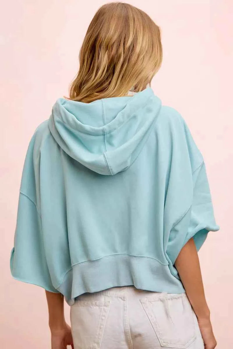 Woman wearing a light blue washed terry short sleeve hoodie top with drawstring hood and relaxed oversized fit by BiBi. Back View