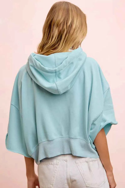 Woman wearing a light blue washed terry short sleeve hoodie top with drawstring hood and relaxed oversized fit by BiBi. Back View
